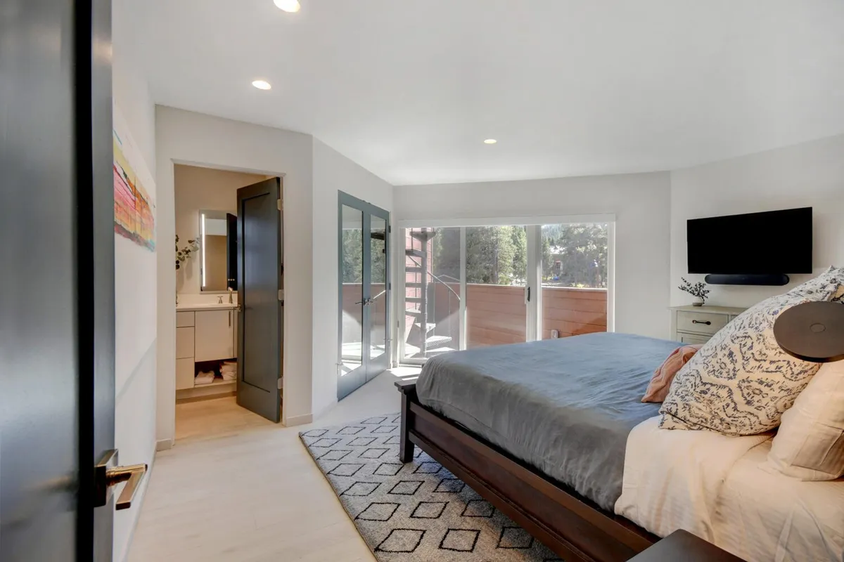 A serene master bedroom featuring a comfortable bed with decorative pillows, large glass doors that open to an outdoor space, and a wall-mounted TV. The room is bathed in natural light, with premium linens giving an inviting, luxury feel.