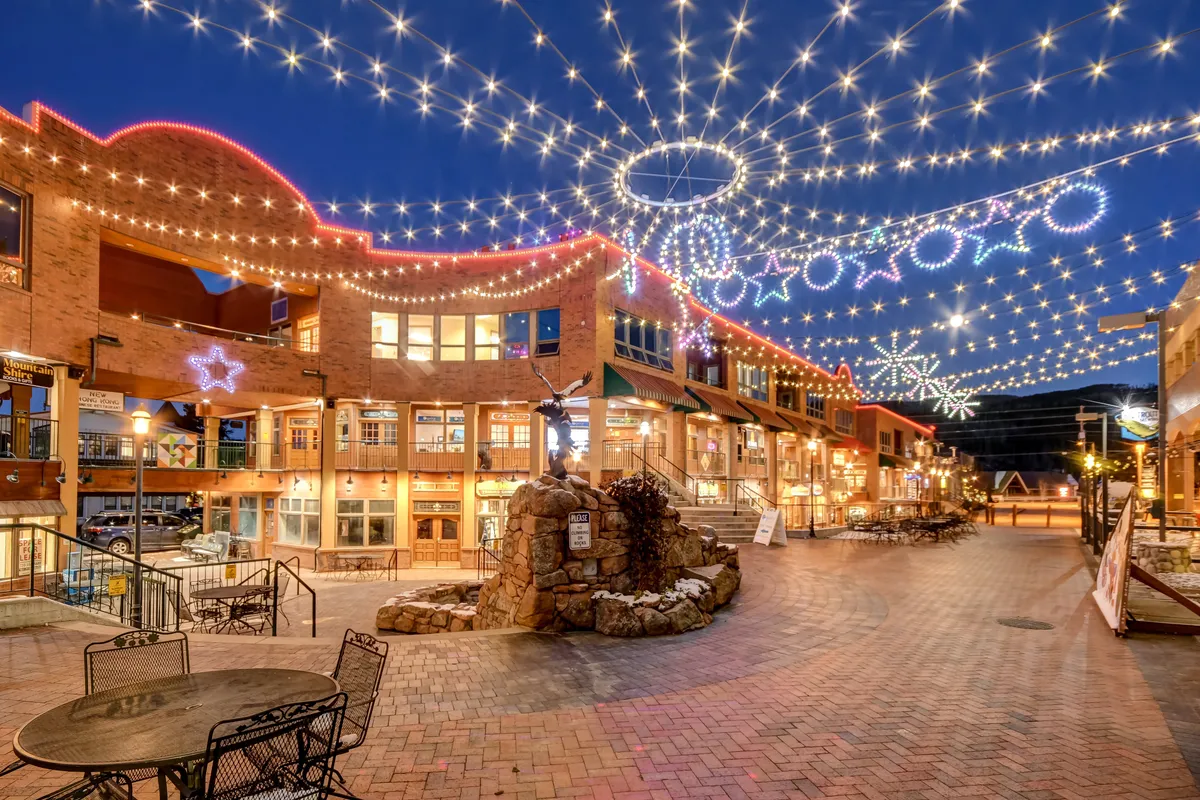 Twinkling decorative lights illuminate a charming plaza with a stone fountain, outdoor seating areas, and inviting storefronts. The atmosphere is festive and social, perfect for evening strolls, dining, or community events.