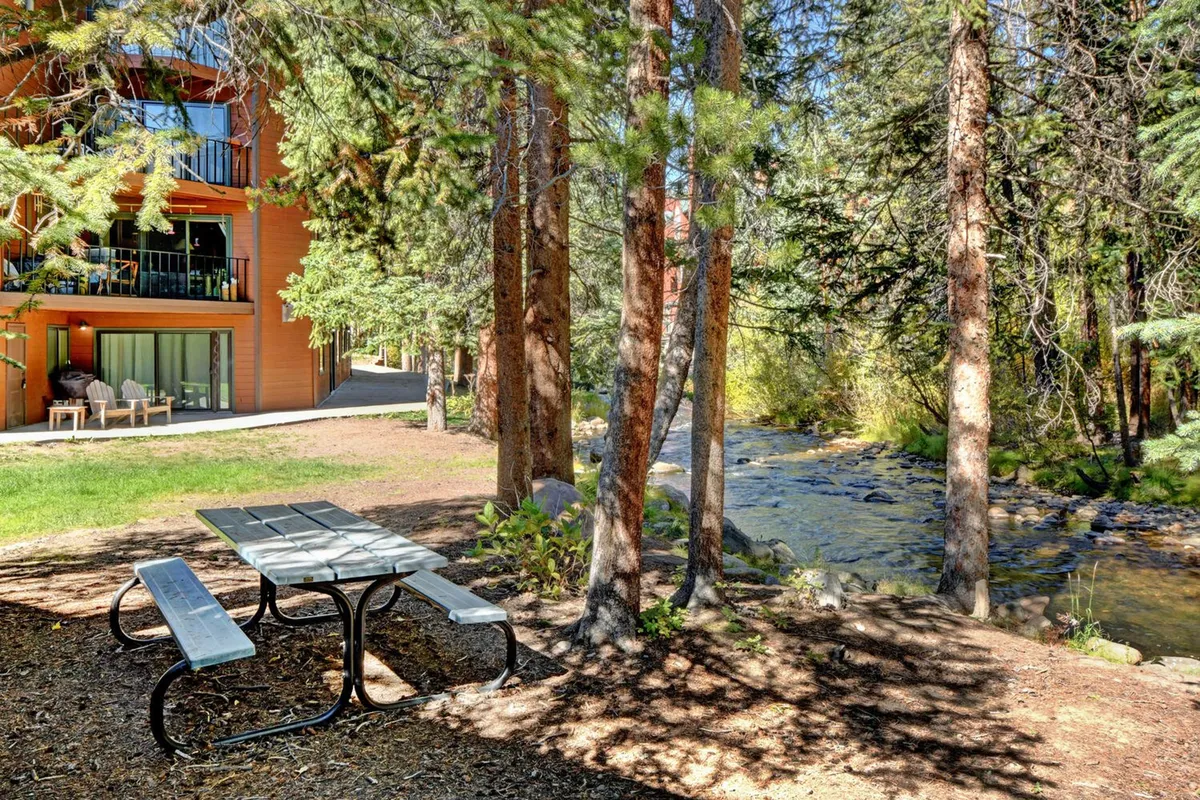 A peaceful outdoor scene featuring a picnic table beside a gentle stream, surrounded by trees, with the balcony of the building visible in the background. The setting invites family gatherings, outdoor meals, and mingling with nature.