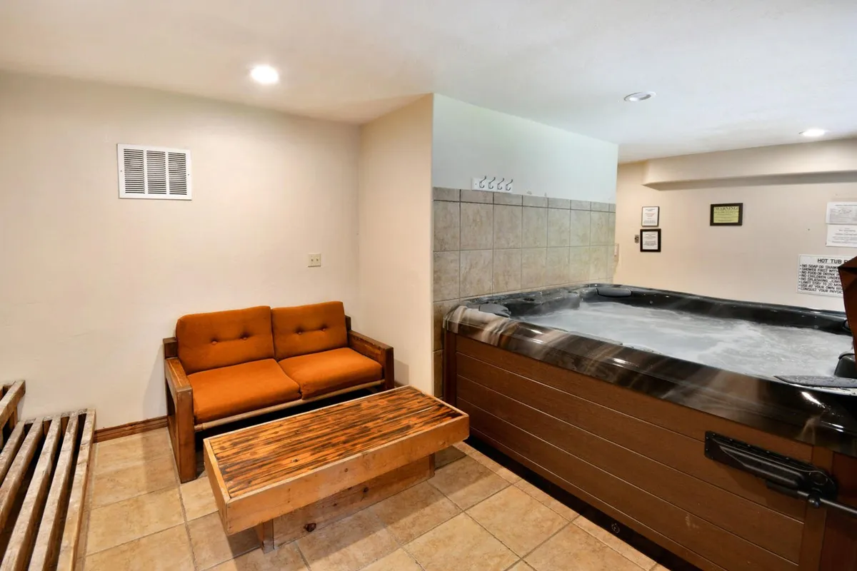 An inviting indoor hot tub area featuring a bubbling hot tub, comfortable orange couch, and a wooden coffee table. The setting exudes relaxation and comfort, perfect for unwinding after a day of adventure.