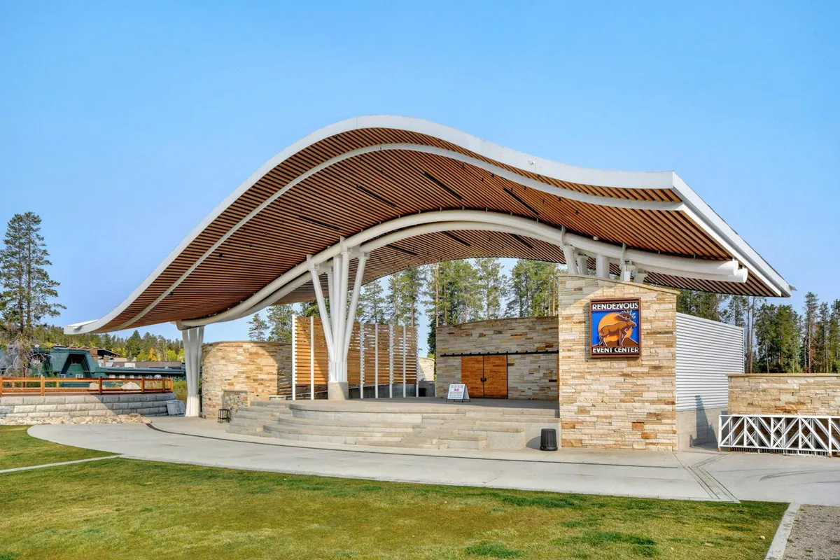 This image captures a lively outdoor stage area framed by a dramatic curved roof structure and a stone facade. The scene is set for community events or celebrations, with a festive atmosphere and plenty of open space for gatherings.