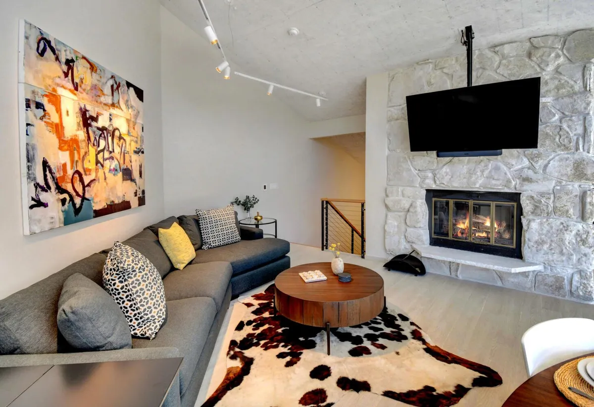 This image captures a spacious great room featuring a comfortable sofa, a stone fireplace, a large abstract painting, and a round coffee table. The seating area is arranged for conversation or relaxation, with warm lighting and cozy mountain-inspired decor.