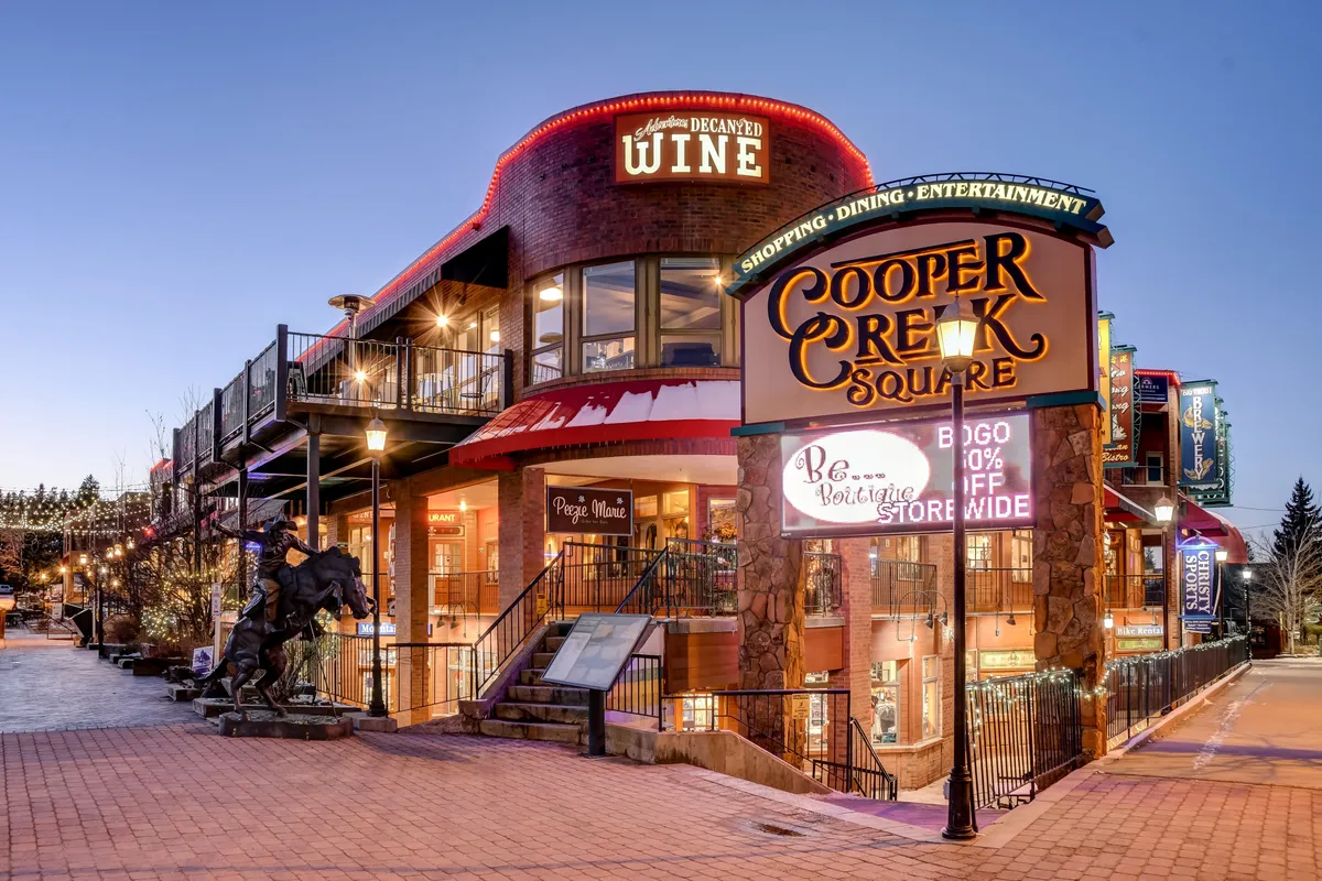 A bustling streetscape with the Cooper Creek Square sign, wine shop, outdoor seating area, staircase leading to an upper level, and decorative lighting. Lively crowds suggest shopping, dining, and entertainment in a festive, community-oriented setting—ideal for après-adventure socializing.