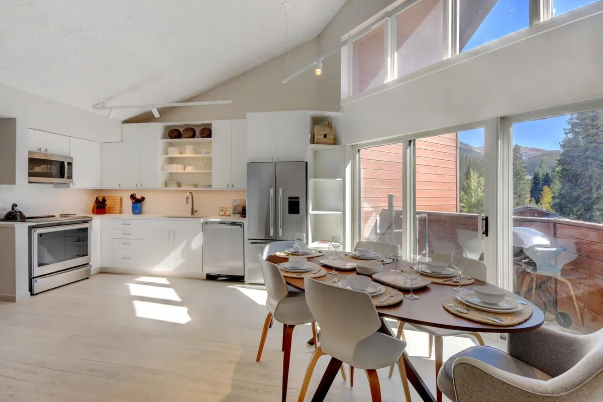 A spacious dining table is set for a meal, surrounded by ample seating and positioned near large windows that let in abundant daylight. The backdrop includes a glimpse of modern kitchen appliances, underscoring a seamless kitchen-dining flow. The mood is cozy and inviting, ideal for family meals and social gatherings.