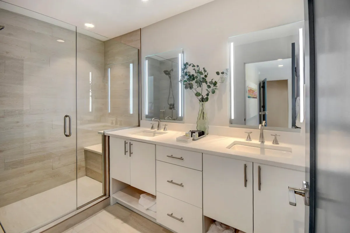 A modern bathroom with double sinks, a glass shower enclosure, and a decorative vase with greenery. The clean lines and tranquil decor evoke a spa atmosphere, emphasizing self-care and relaxation.