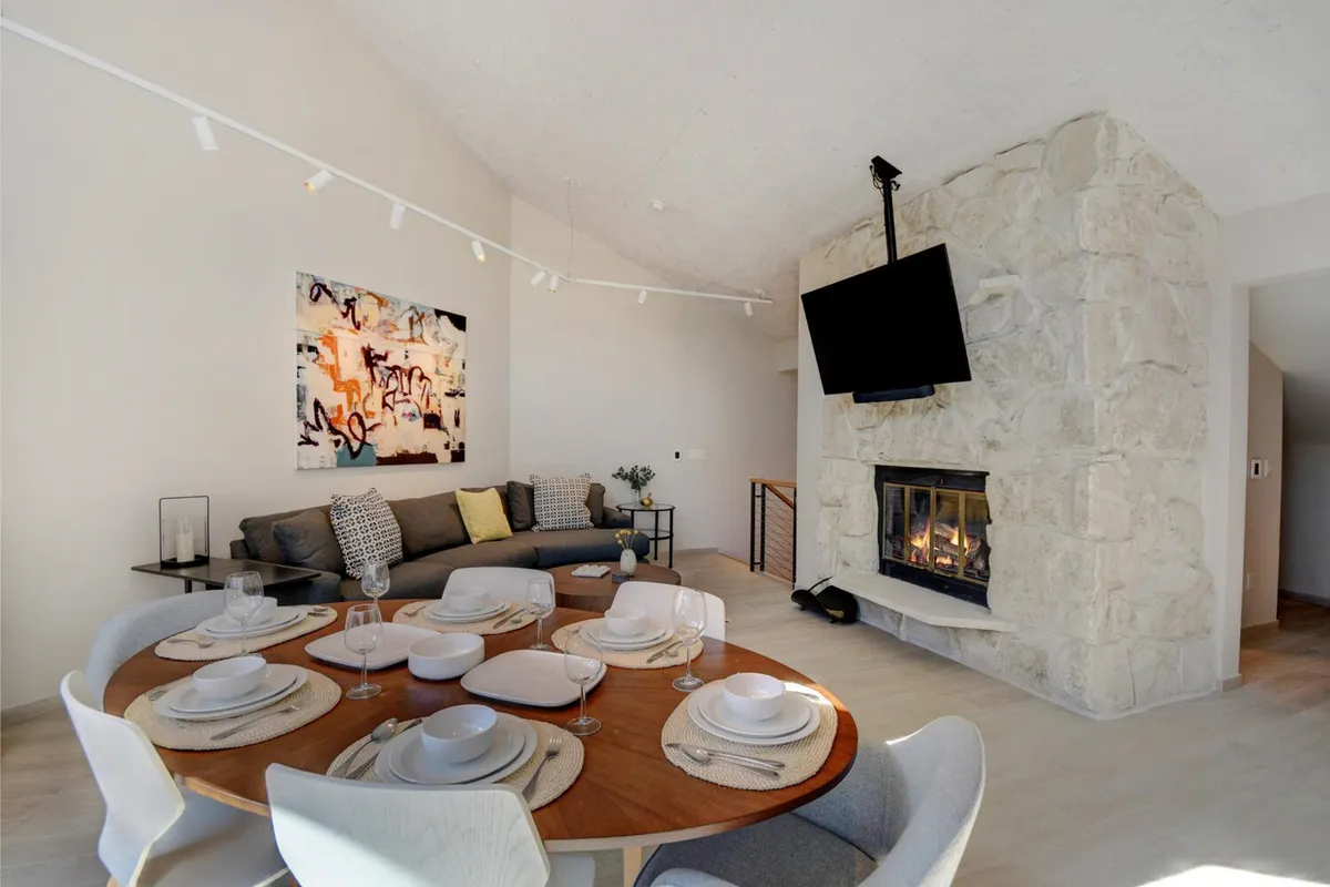 The photo shows a warmly lit living/dining area featuring a dining table, a fireplace, modern artwork, and a sofa, all arranged in an open, social layout. The ambiance is both contemporary and cozy, ideal for hosting gatherings or enjoying board games.